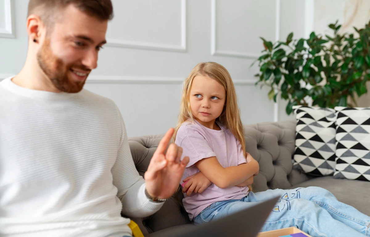 Батько розмовляє з донькою на дивані й жестом показує межу або правило, поки дитина реагує стримано й недовірливо