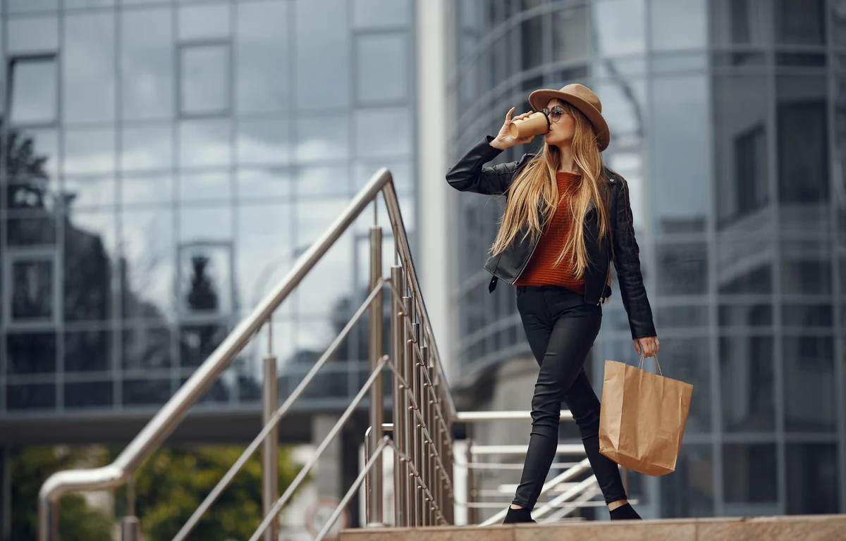 Woman in a hat by a modern business center - city casual as a response to the era of overload: restrained style, comfortable pieces, and conscious clothing choices without chasing trends.