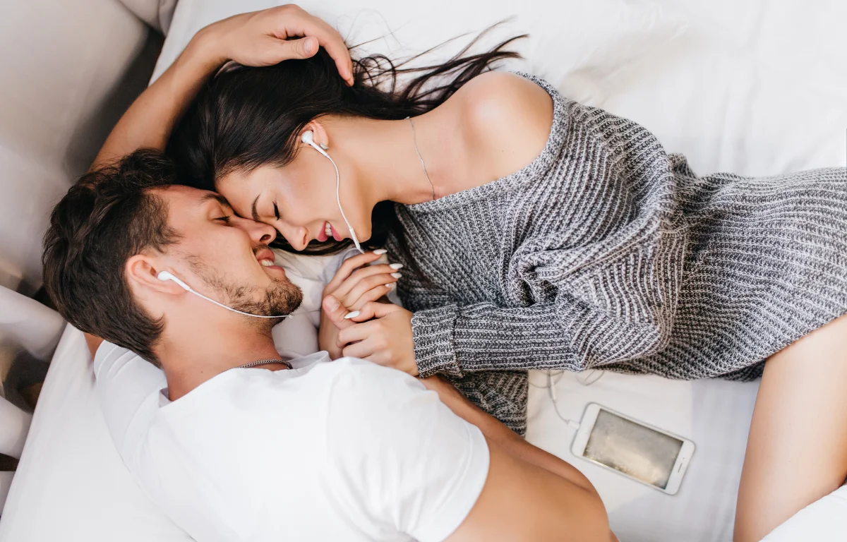 Couple lying on a bed with earphones, holding hands in a moment of modern intimacy