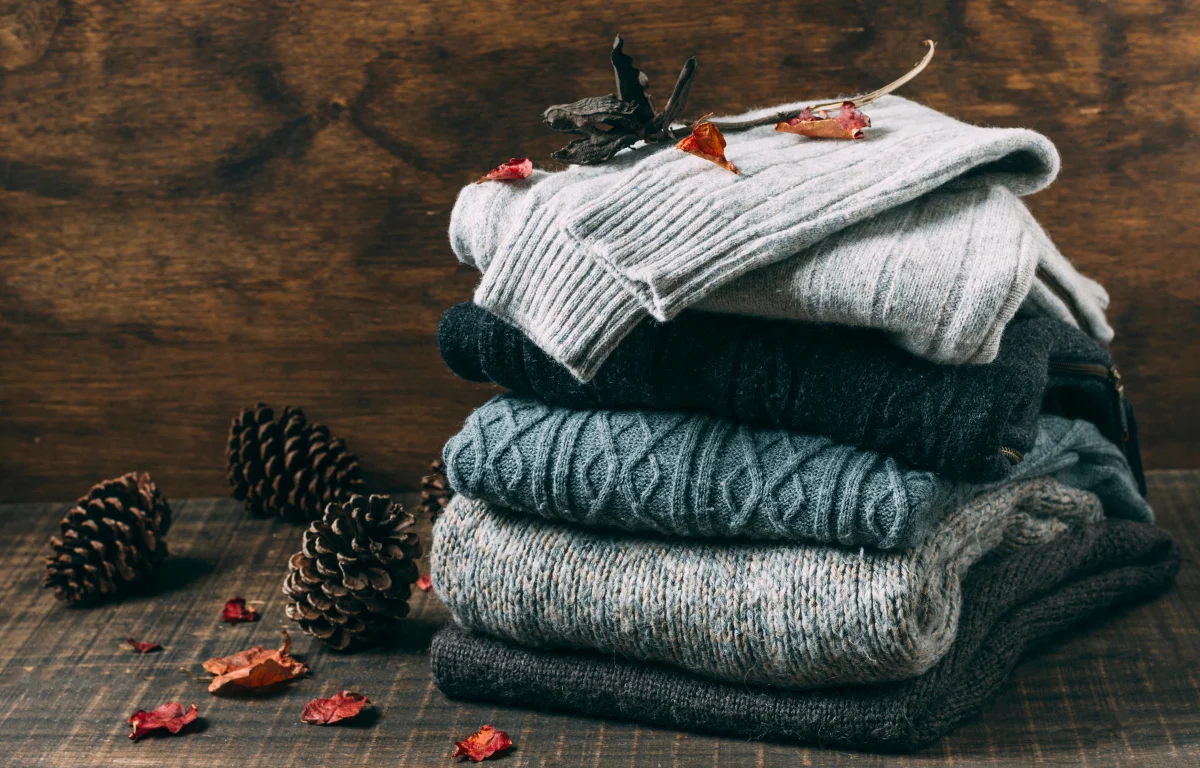 Folded wool sweaters with textured knits arranged on wood beside pinecones
